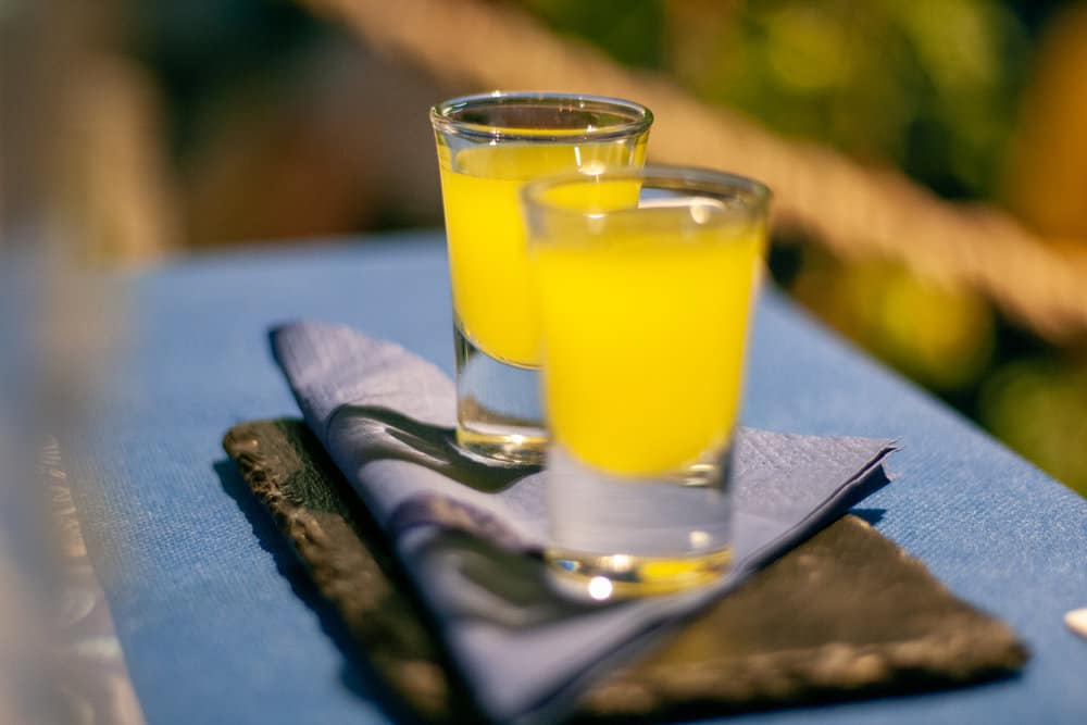 Two shot glasses filled with bright yellow liquid sit on a blue napkin atop a slate surface.