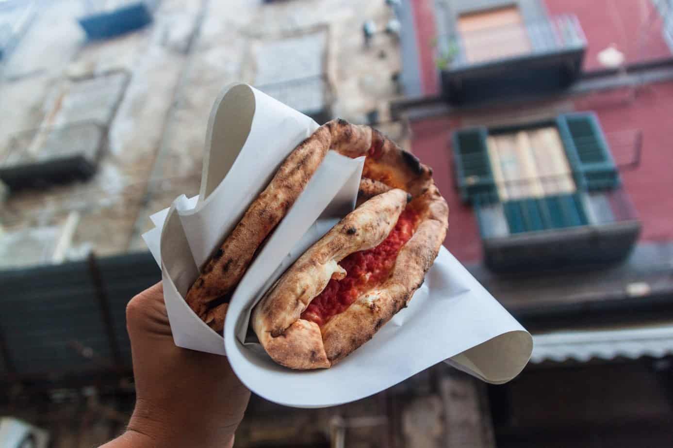 A hand holds two folded pizzas wrapped in paper, with blurred buildings in the background.