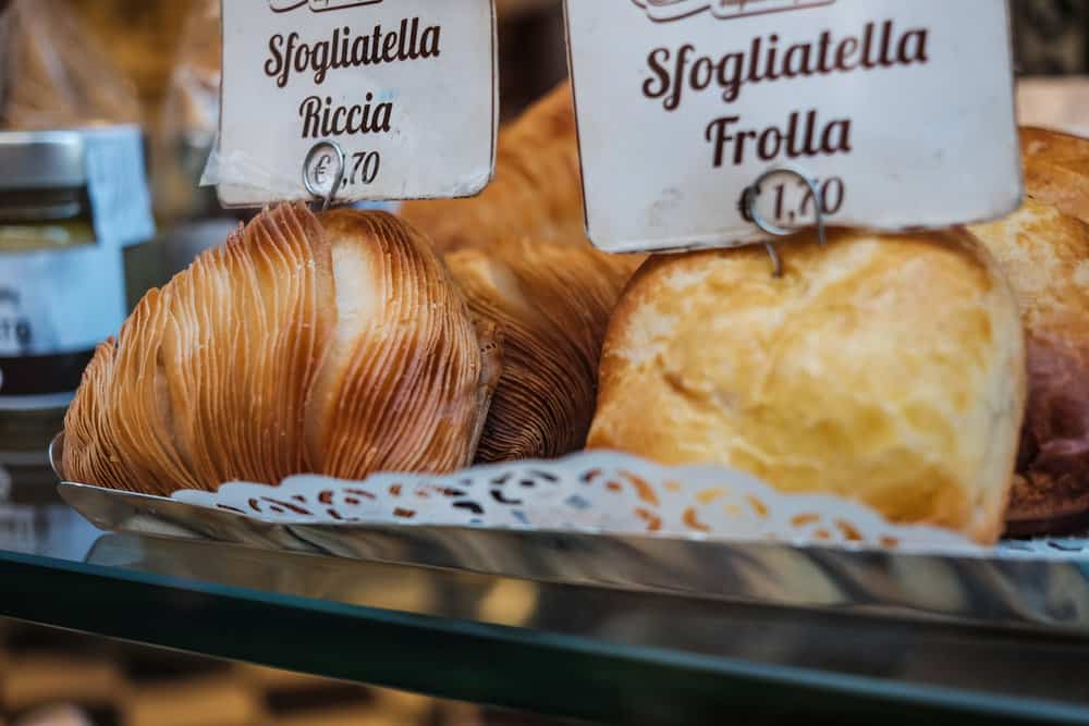Sfogliatella pastries in Naples Italy, elegantly arranged on a lace doily, are labeled "Riccia" and "Frolla," with prices of 1.70 and 1.40 euros respectively.