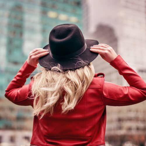 Person with long blonde hair wearing a black hat and red jacket, standing in front of tall buildings in a cityscape.