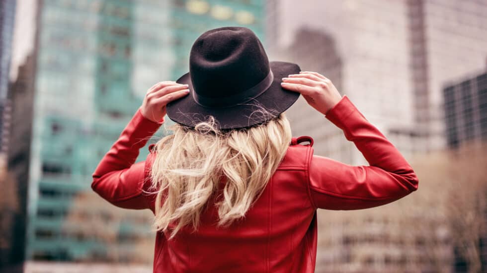 Person with long blonde hair wearing a black hat and red jacket, standing in front of tall buildings in a cityscape.