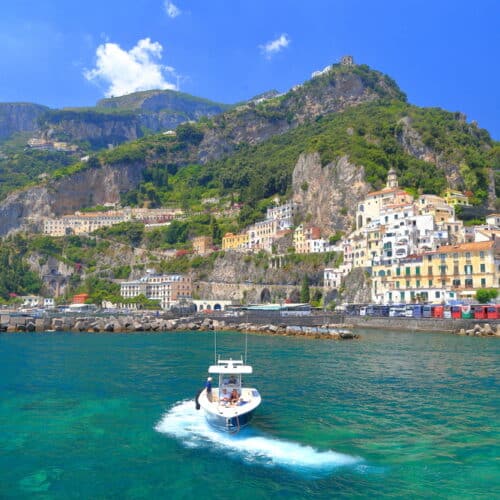 A small boat glides through the turquoise waters of Amalfi Harbour, with a hillside town and lush greenery in the background under a clear blue sky.