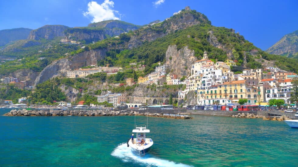 A small boat glides through the turquoise waters of Amalfi Harbour, with a hillside town and lush greenery in the background under a clear blue sky.