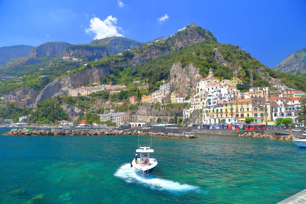 A small boat glides through the turquoise waters of Amalfi Harbour, with a hillside town and lush greenery in the background under a clear blue sky.
