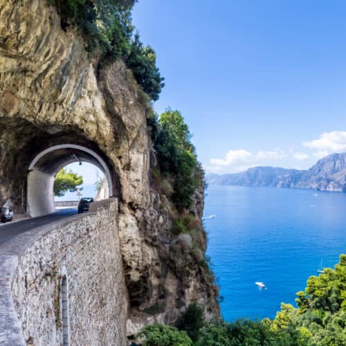 A coastal road with a tunnel hugs a cliffside on the Amalfi Coast, overlooking a clear blue sea and distant mountains under a bright sky.