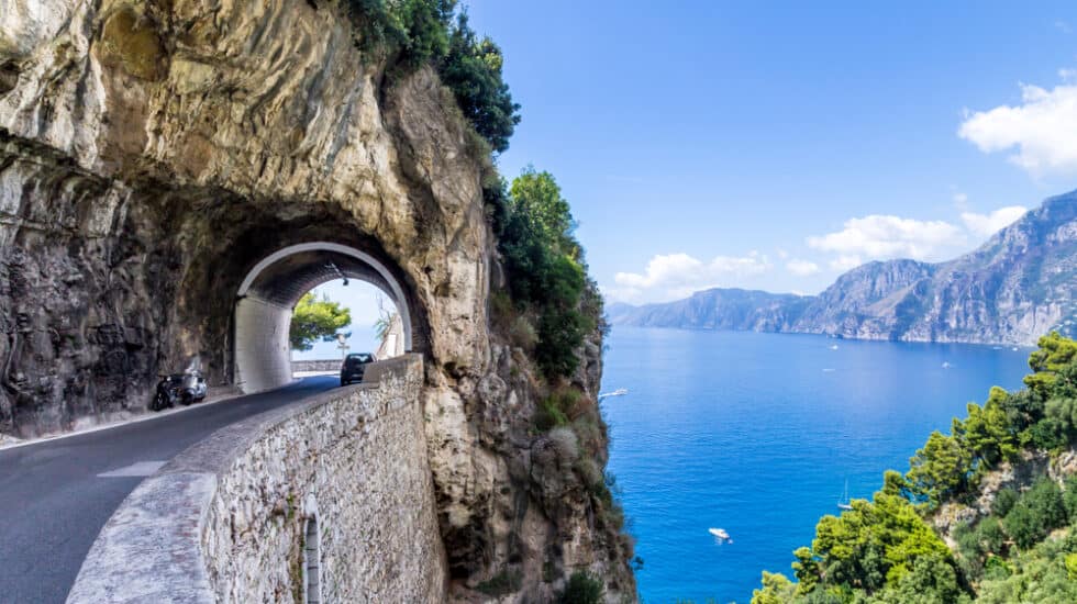A coastal road with a tunnel hugs a cliffside on the Amalfi Coast, overlooking a clear blue sea and distant mountains under a bright sky.