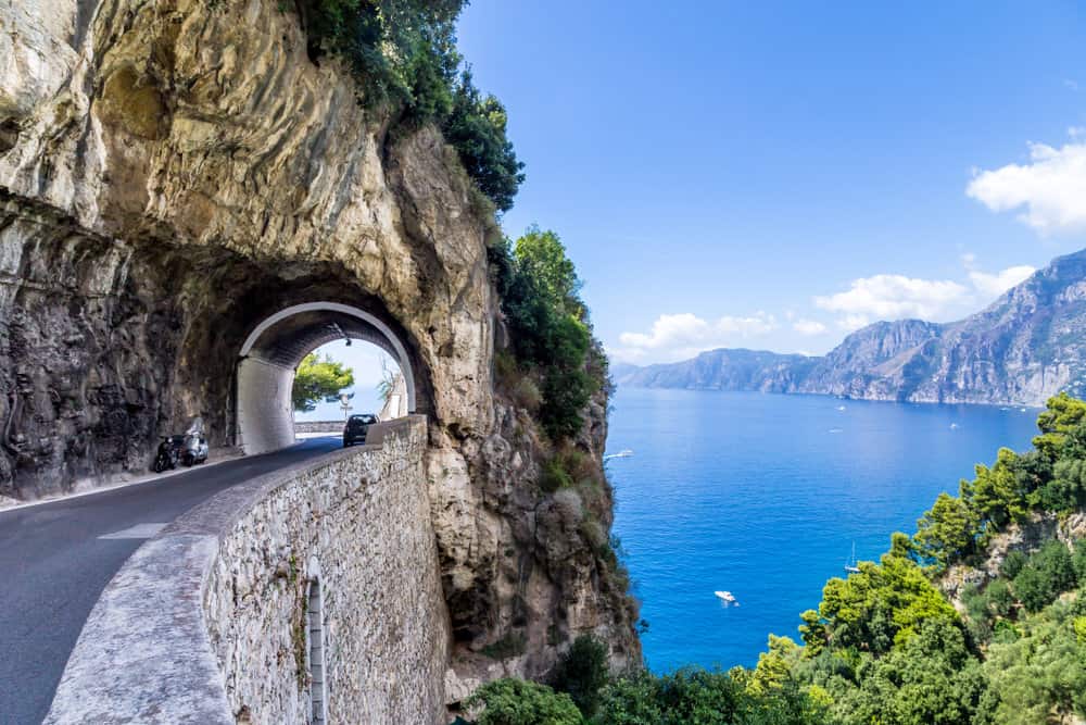A coastal road with a tunnel hugs a cliffside on the Amalfi Coast, overlooking a clear blue sea and distant mountains under a bright sky.