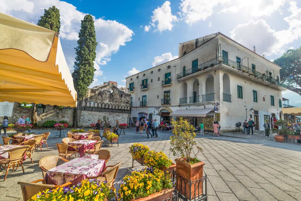 Outdoor café with tables and chairs on a sunny plaza in Duomo Square, surrounded by flowers. A few people stroll near a historic white building with balconies, adding to the charm of this iconic spot.