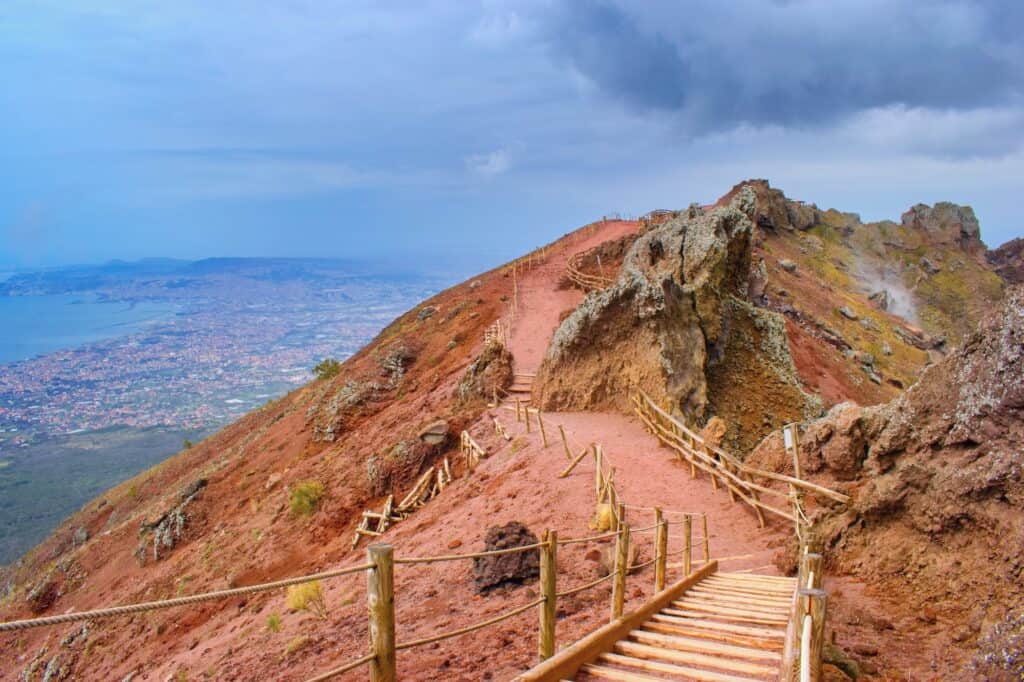 A wooden crater path at Mount Vesuvius winds along a rocky mountain slope, offering a breathtaking view of the city by the sea under a cloudy sky.