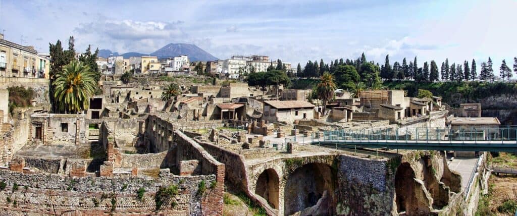 The ancient ruins of Herculaneum showcase partially intact buildings surrounded by lush palm trees, all under the watchful gaze of Mount Vesuvius against a backdrop of clear skies.