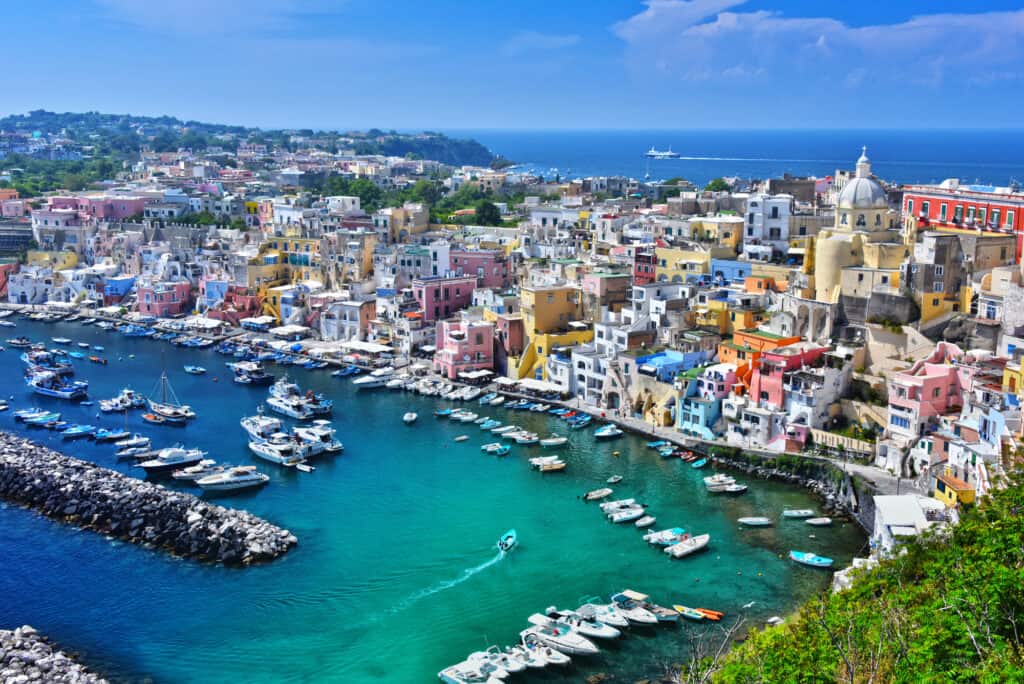 The vibrant coastal town of Procida Island near Naples showcases numerous boats docked in a clear blue harbor, surrounded by colorful buildings and lush greenery under the expansive blue sky.