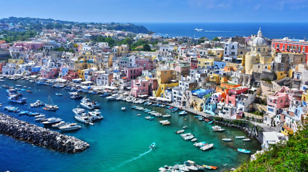 The vibrant coastal town of Procida Island near Naples showcases numerous boats docked in a clear blue harbor, surrounded by colorful buildings and lush greenery under the expansive blue sky.