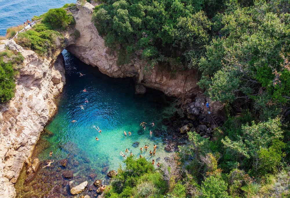 Aerial view of Regina Giovanna's natural coastal lagoon, resembling a queen's bath, surrounded by lush greenery, with people swimming in its clear blue waters.