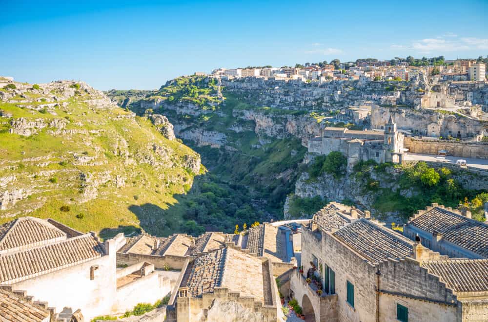From the Musma Museum at Palazzo Pomarici, enjoy a breathtaking view of Matera's rocky landscape adorned with historic stone buildings and a deep valley, all framed under a clear blue sky.