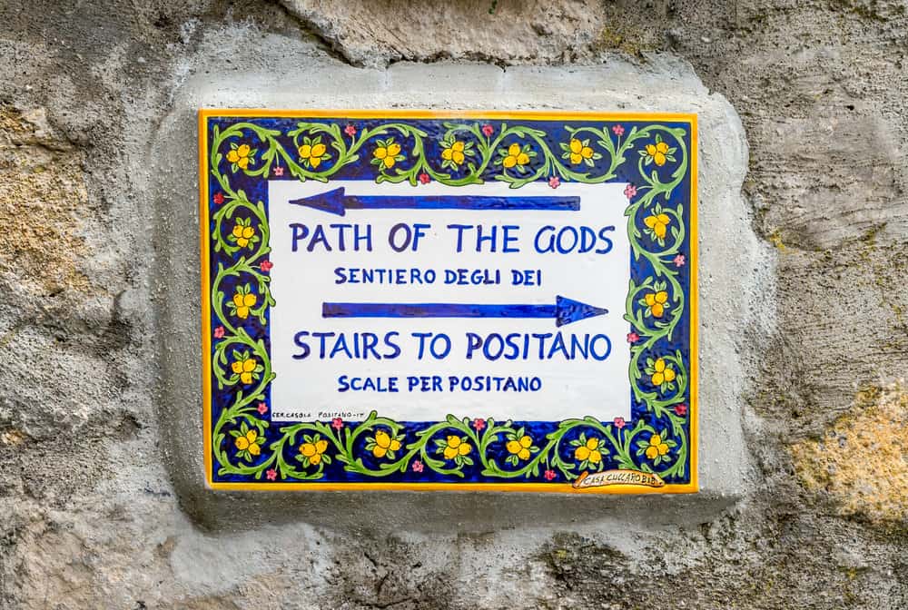 A decorative sign on a stone wall indicates directions: left to "Path of the Gods" and right to "Stairs to Positano," with text in English and Italian.