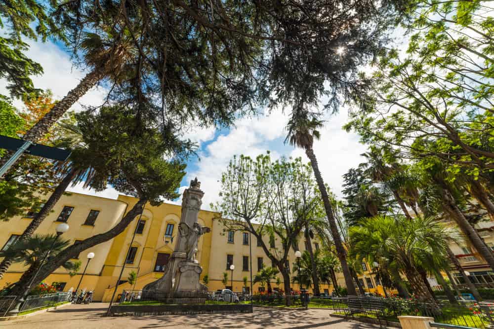 A sunny day in Piazza della Vittoria, where a statue stands amid benches, framed by tall palm trees and a yellow building in the background.