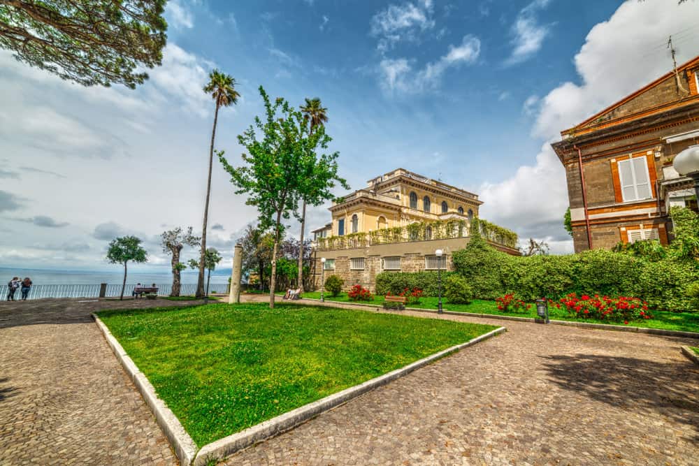A cobblestone pathway surrounds a lush green lawn with trees and flowers, leading to the stately Villa Comunale di Sorrento, standing proudly under a partly cloudy sky.
