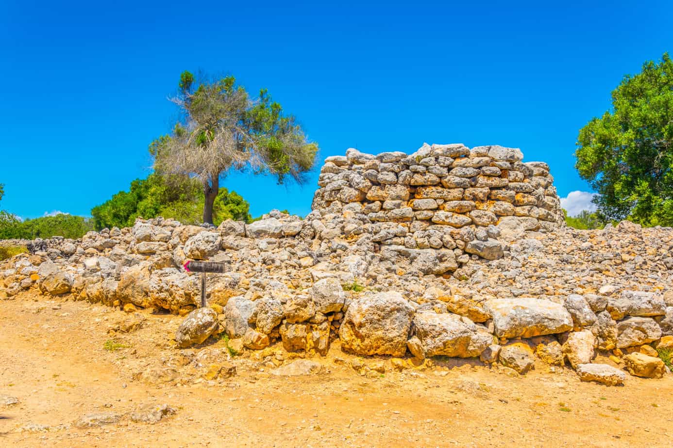 Under a clear blue sky, the ancient stone structure of the Ruins of Talayot Capocorb Vell rises majestically, surrounded by trees and dry ground. A small signpost stands nearby, silently narrating tales of history long past.