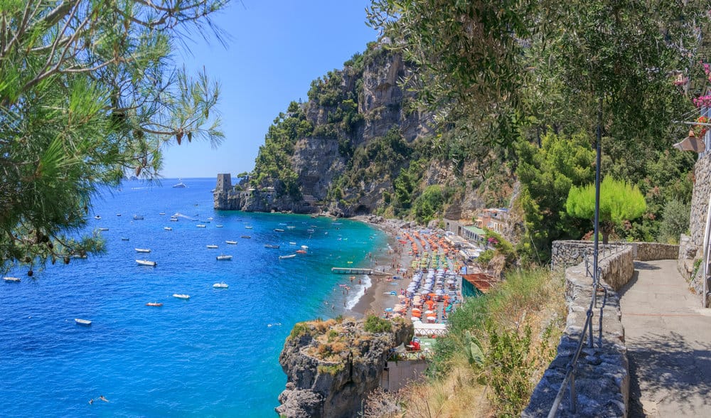 Coastal view of Fornillo Beach with clear blue water, small boats, a rocky shoreline, vibrant beach umbrellas, and cliffside vegetation under a clear sky.