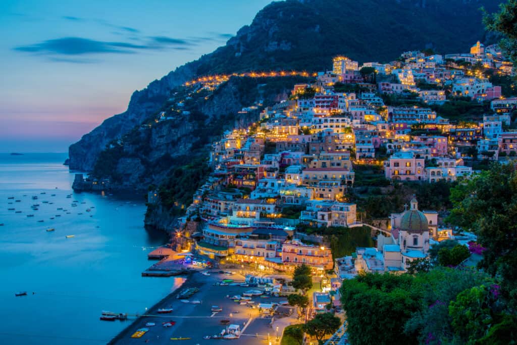 The coastal town of Positano, Italy is illuminated by colorful lights at dusk, with a calm sea and boats in the water, set against a mountainous backdrop.