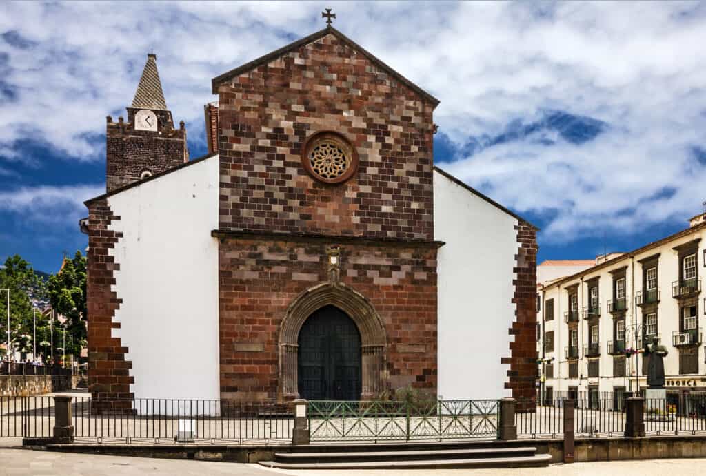 A historic stone church, reminiscent of the Funchal Cathedral, features a towering clock against a blue sky. It has a large arched entrance with a circular window above, inviting visitors to explore its timeless beauty.