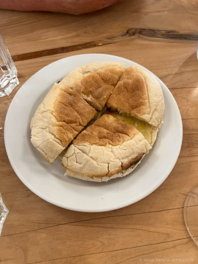 A toasted sandwich, made with the traditional Bolo do caco bread, is cut into quarters and placed on a white plate atop a wooden table.