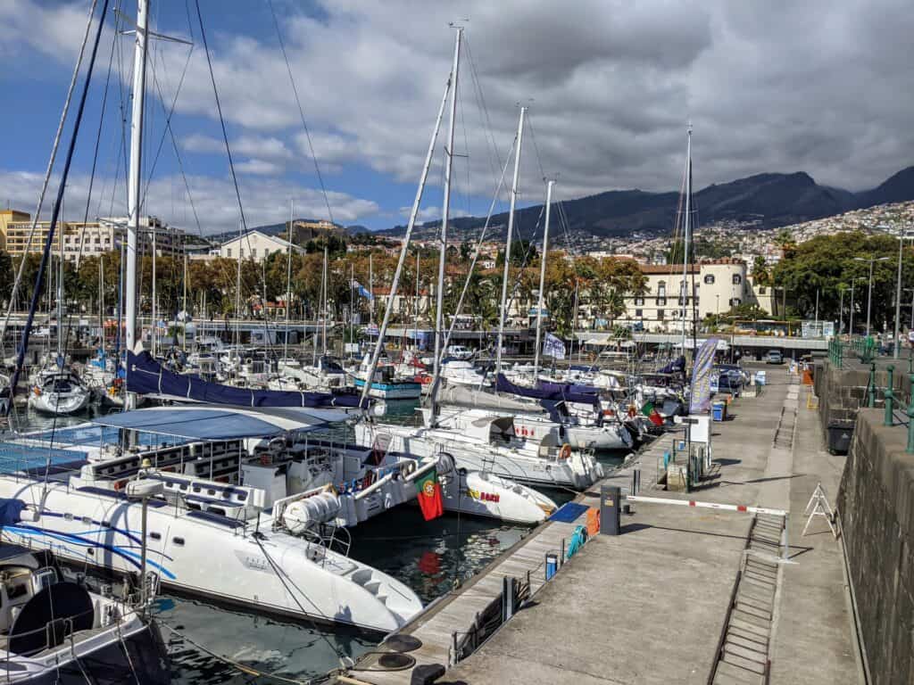 Several sailboats and catamarans docked at a marina with mountains and buildings in the background under a partly cloudy sky.