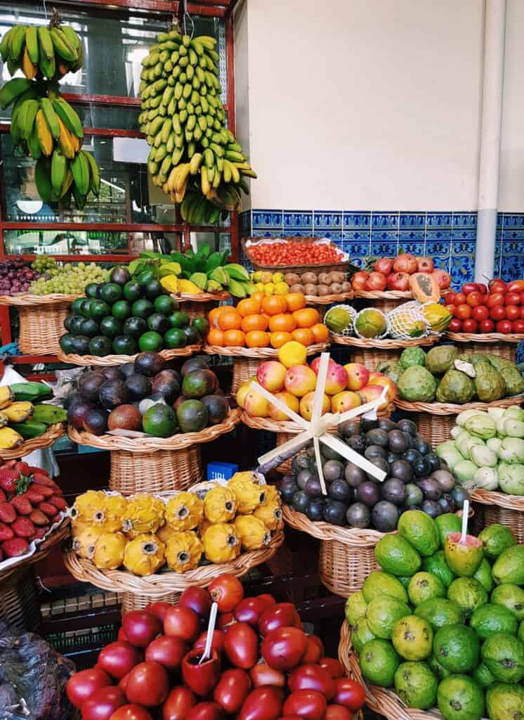 A vibrant fruit market display reminiscent of Mercado do Lavradores features baskets of bananas, oranges, apples, avocados, and other tropical fruits artfully arranged on shelves.