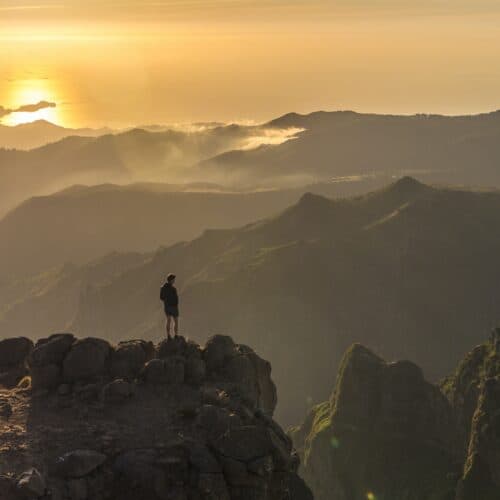 Perched on the rocky cliff of Pico Do Arieiro, a person gazes out at the misty mountains painted in hues of sunset.