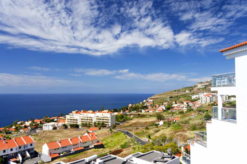 Scenic coastal view of Canico de Baixo resort with charming red-roofed buildings nestled among lush greenery, and the vast ocean stretching out under a blue sky with scattered clouds.