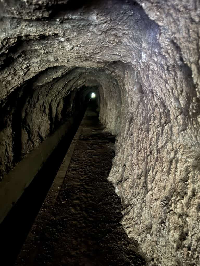 A dimly lit rocky tunnel with uneven walls and dark flooring extends forward, on the levada Nova trail, with a faint light visible at the end.
