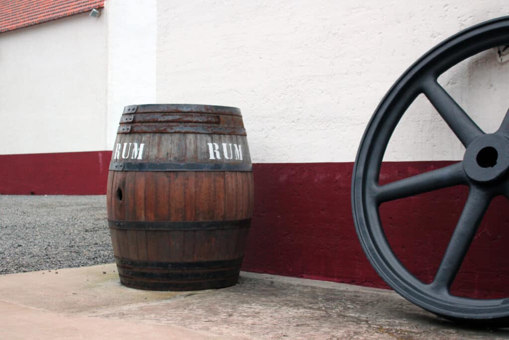 A wooden barrel labeled "RUM" stands next to a large, black wheel by a building with white and red walls.