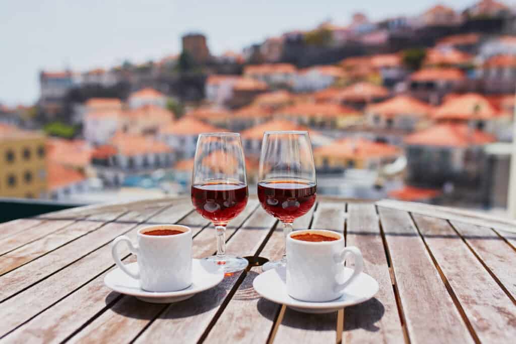 Two cups of coffee and two glasses of red wine on a wooden table, with a blurred hillside town in the background.