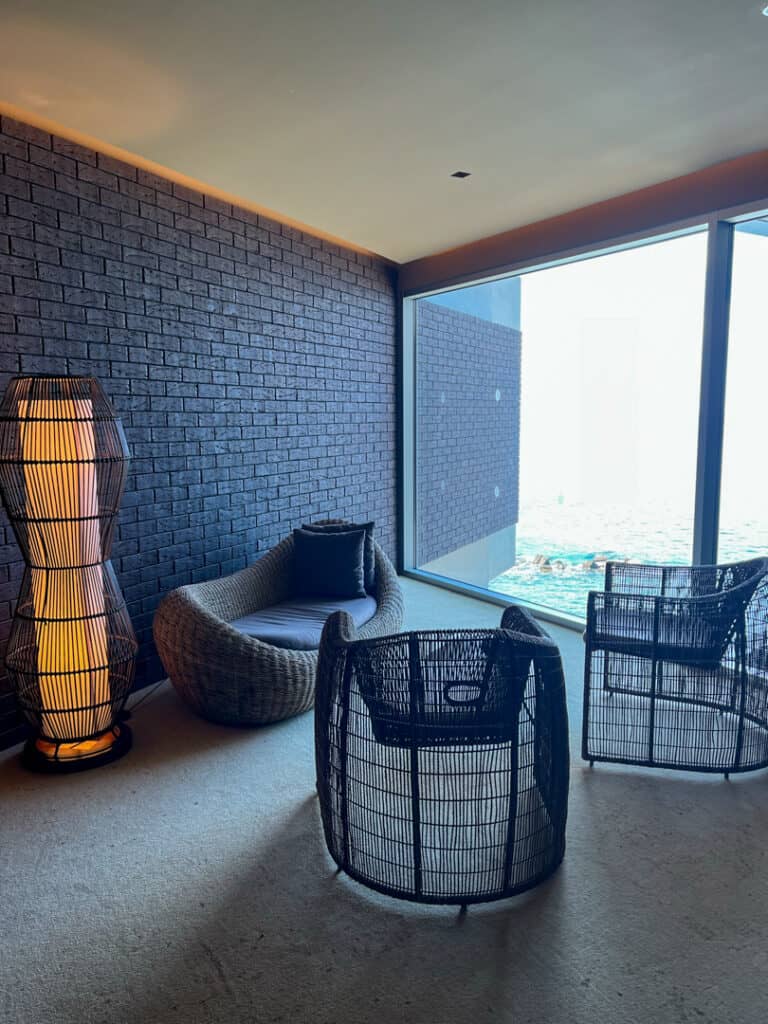 A cozy room with a wicker sofa, two wicker chairs, a tall floor lamp, and a large window overlooking a cityscape. The walls feature a textured brick design.