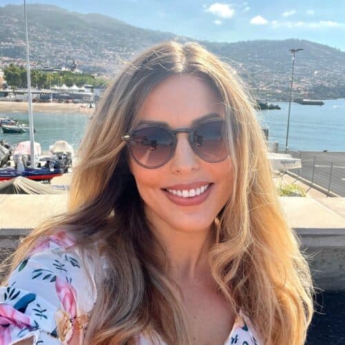 A person with long hair and sunglasses smiles in front of a waterfront with boats, under a clear blue sky in Funchal, capturing the essence of their Madeira itinerary.