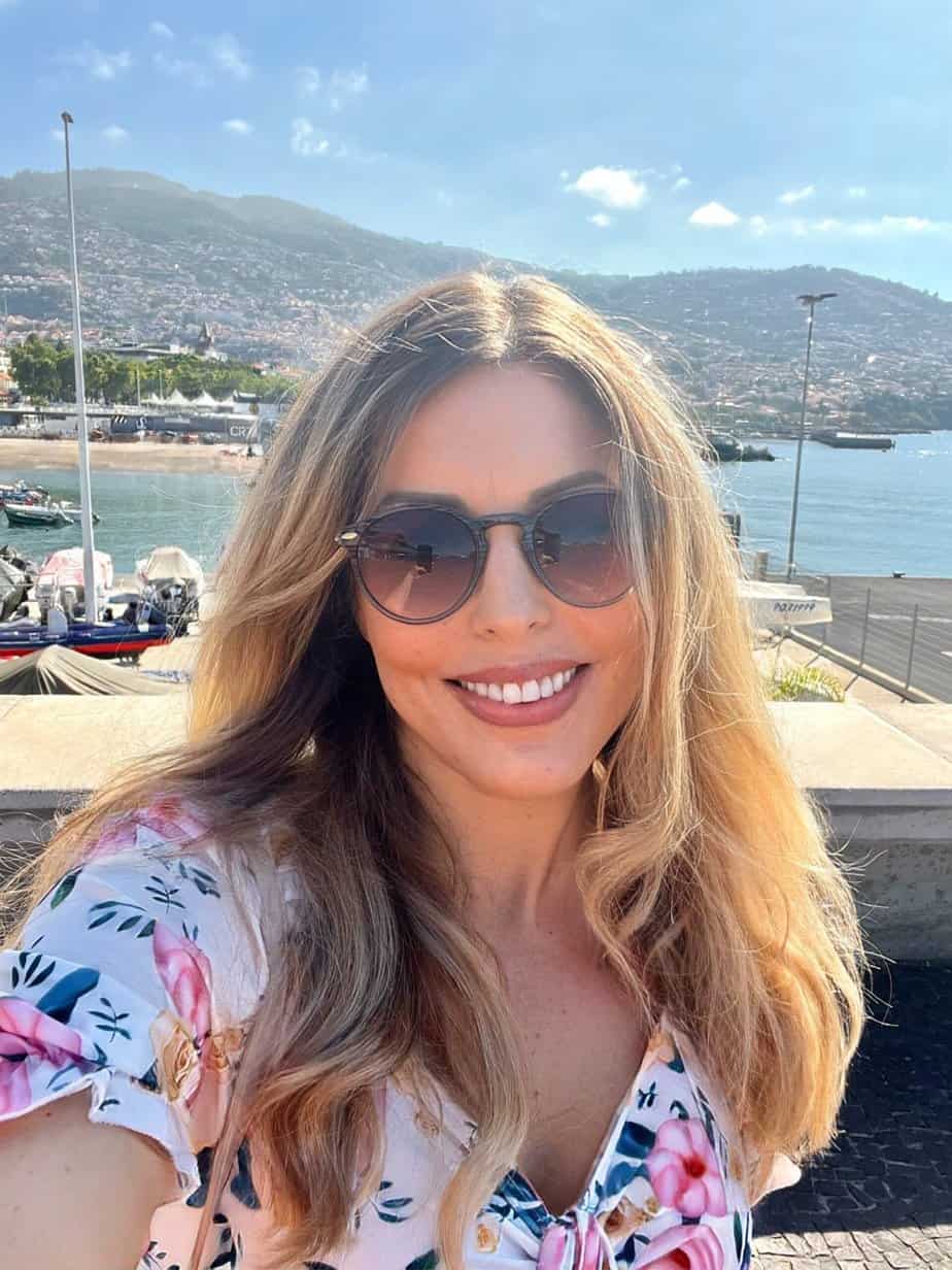 A person with long hair and sunglasses smiles in front of a waterfront with boats, under a clear blue sky in Funchal, capturing the essence of their Madeira itinerary.