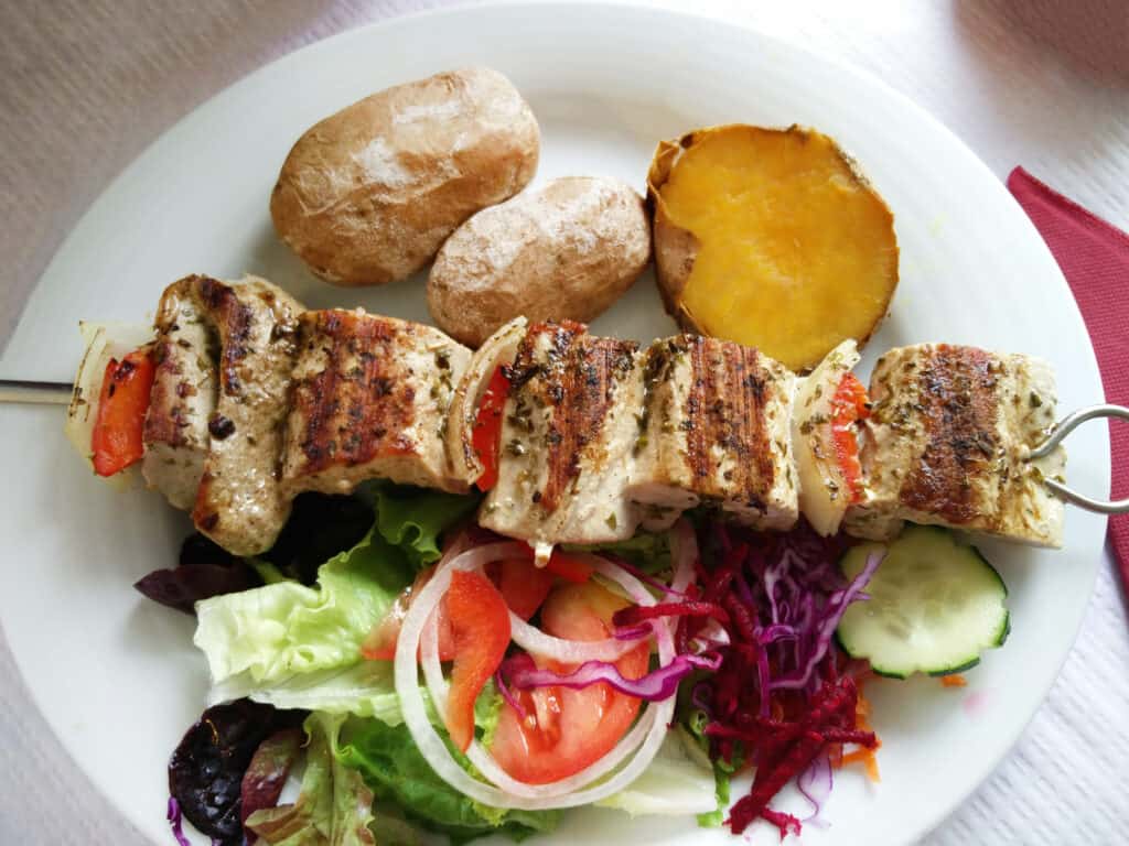 A plate with pork espetada skewers, a baked potato, a slice of roasted sweet potato, and a mixed salad of lettuce, tomato, cabbage, and cucumber.