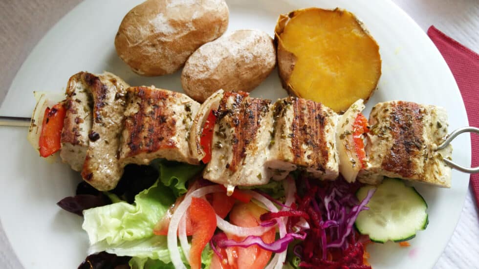 A plate with pork espetada skewers, a baked potato, a slice of roasted sweet potato, and a mixed salad of lettuce, tomato, cabbage, and cucumber.