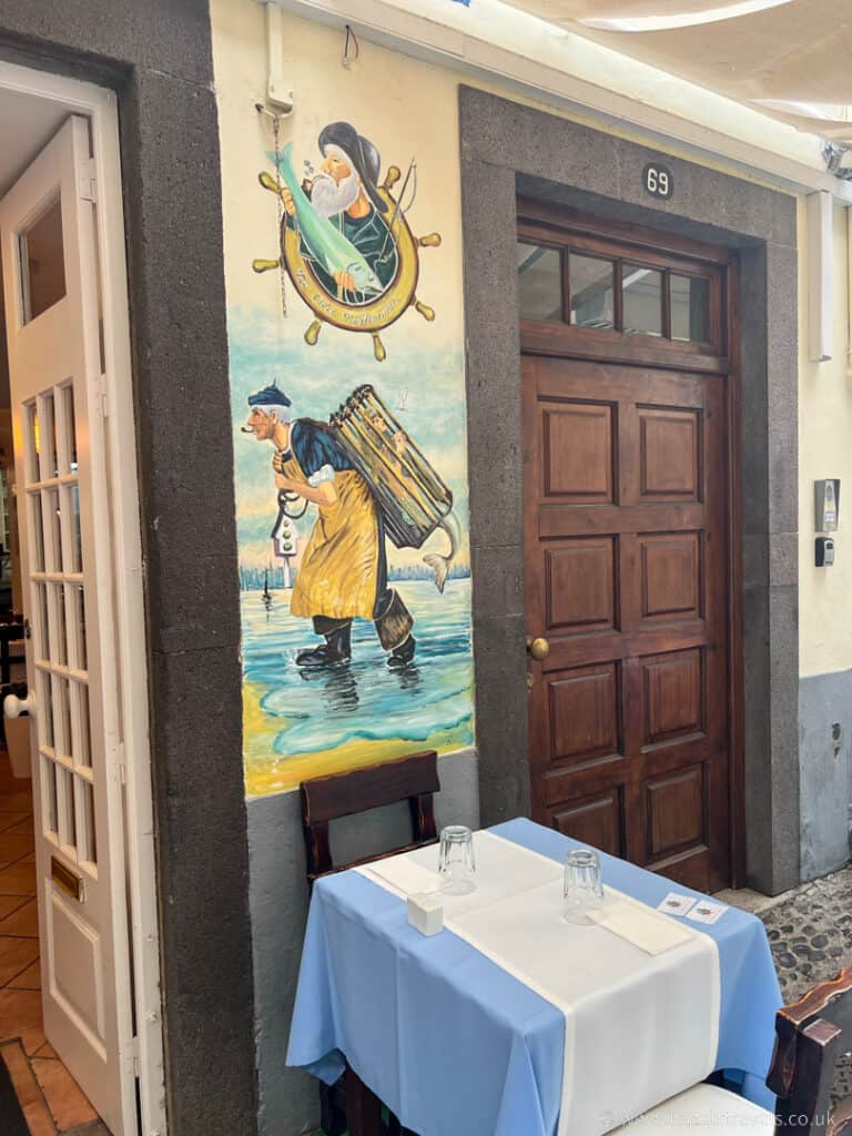 Restaurant entrance with wooden door and mural of a sailor carrying a cask. Two tables with blue tablecloths and glasses are set outside.