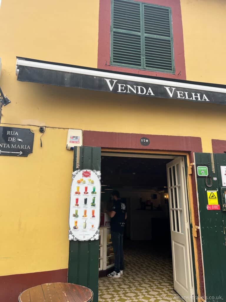 A small café with a yellow and red façade on Santa Maria Street in Funchal, proudly displaying "Venda Velha." Inside, a person stands close to a menu board featuring an array of colorful drinks.