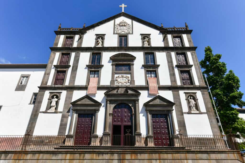 The facade of the Church of Saint John the Evangelist in Funchal features ornate statues, three large wooden doors, and a cross on top, all set against a clear blue sky.