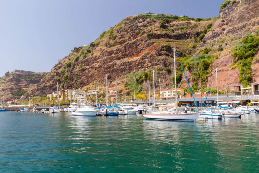 Sailboats and yachts gently anchor in Calheta Marina, nestled against a rocky hillside with charming buildings dotting the background under a clear sky.