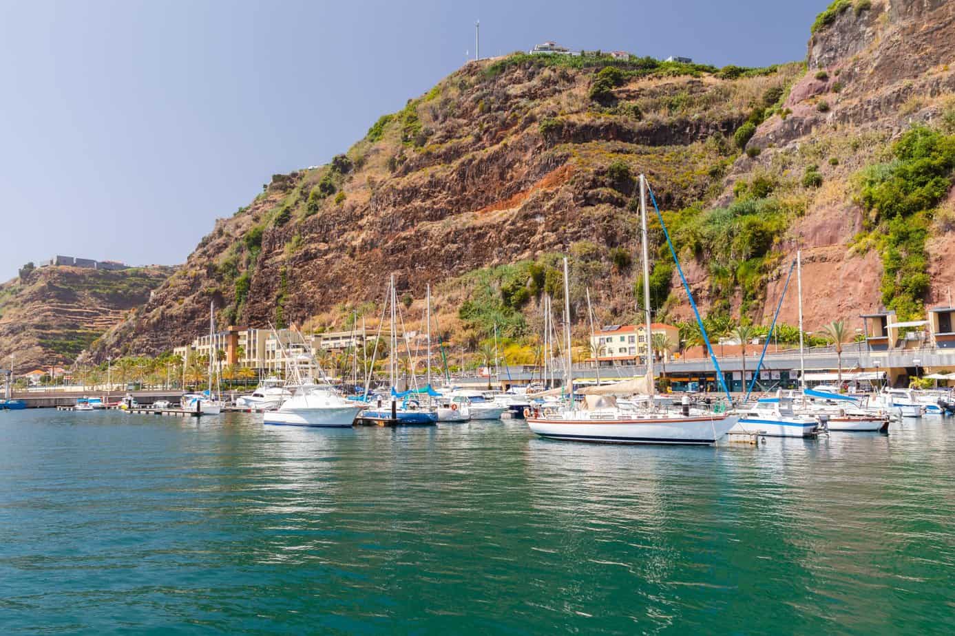 Sailboats and yachts gently anchor in Calheta Marina, nestled against a rocky hillside with charming buildings dotting the background under a clear sky.