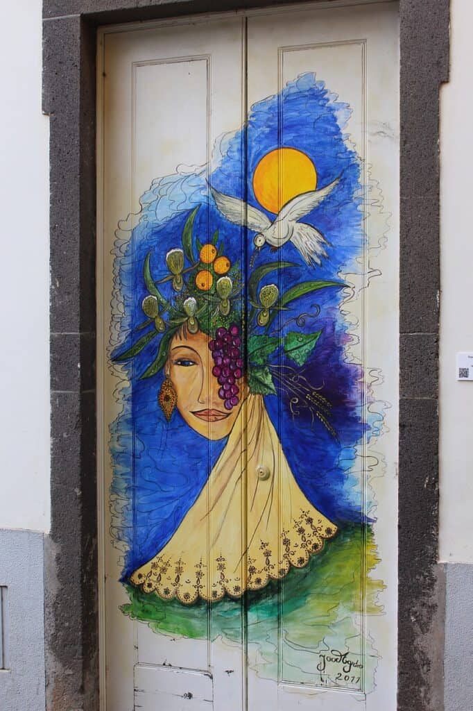 Colorful mural on a door depicting a face with a headdress of fruits, flowers, and leaves, a white bird in flight, and a yellow sun against a blue background.