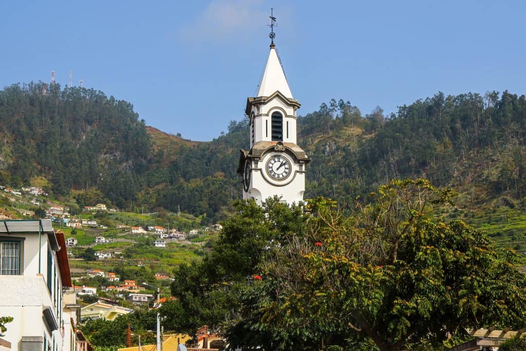 White clock tower with a pointed roof against a backdrop of green hills in Porto Moniz Madeira.