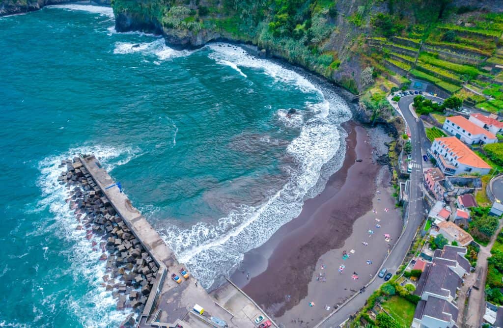 Aerial view of a curved beach with dark sand beside a cliff, with a breakwater and buildings nearby. Seixal, near Porto Moniz in Madeira.