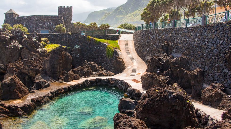 Natural rock pool with a staircase leading up to a stone tower, surrounded by lush hills.