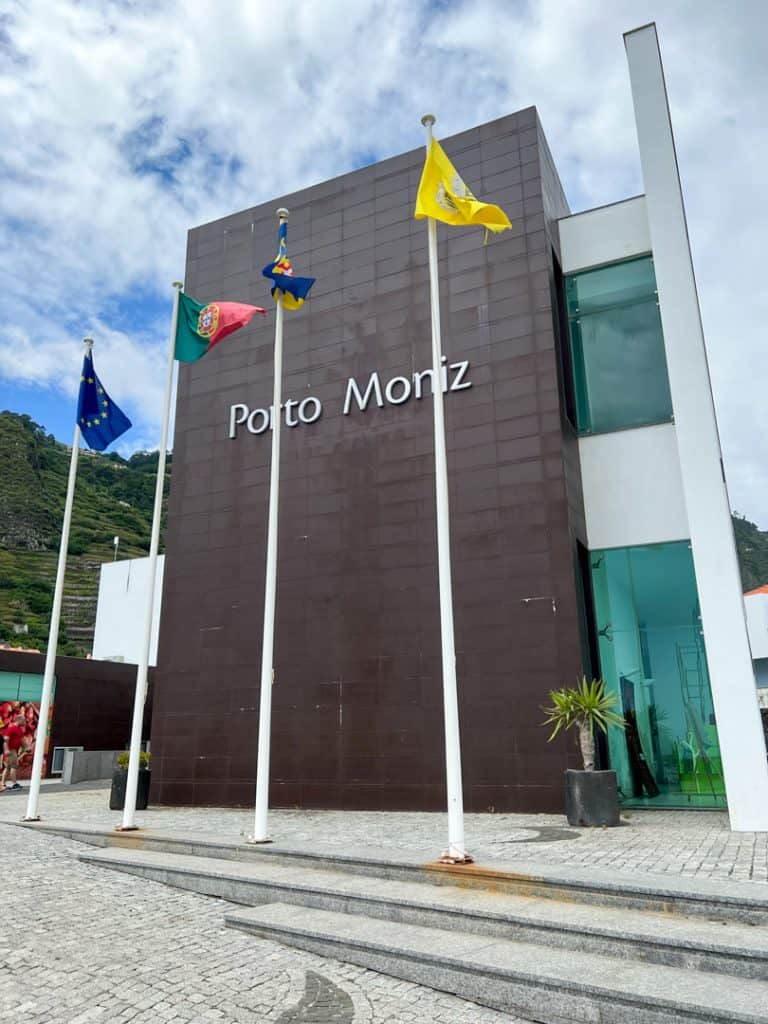 The entrance of a modern building with "porto moniz" written on the facade, flanked by the flags of portugal, the european union, and two other flags under a blue sky with clouds.