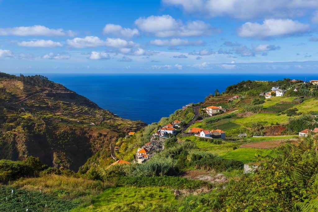A scenic coastal landscape featuring terraced fields, a village, and the ocean horizon under a blue sky with scattered clouds, near Porto Moniz Madeira.