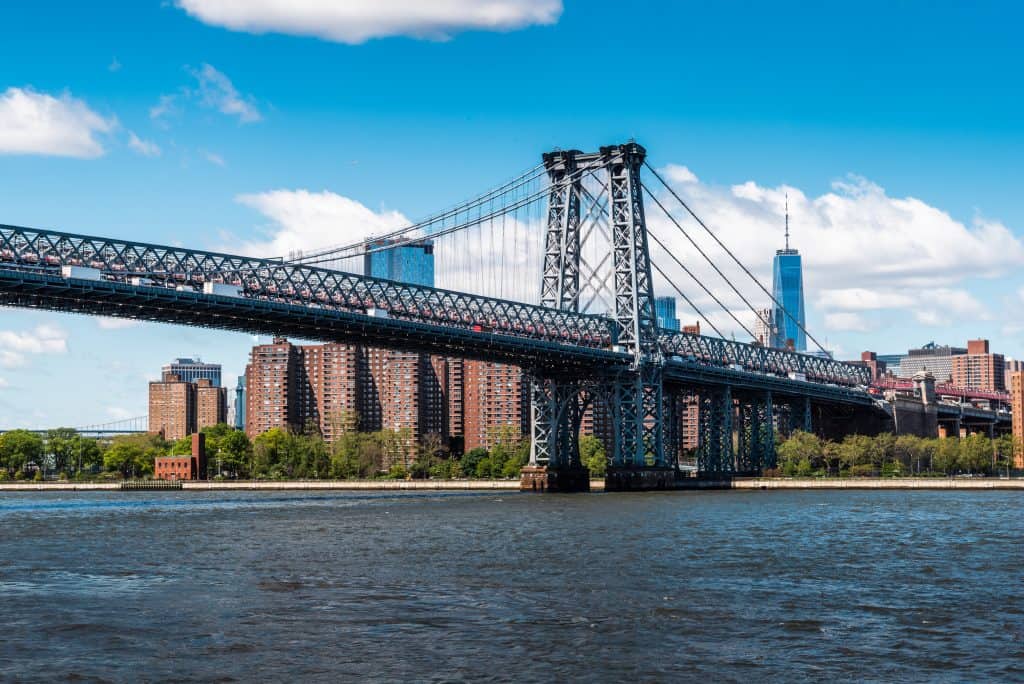 View of a suspension bridge over a river with city skyline in the background on a clear day. Famous bridges in New York.